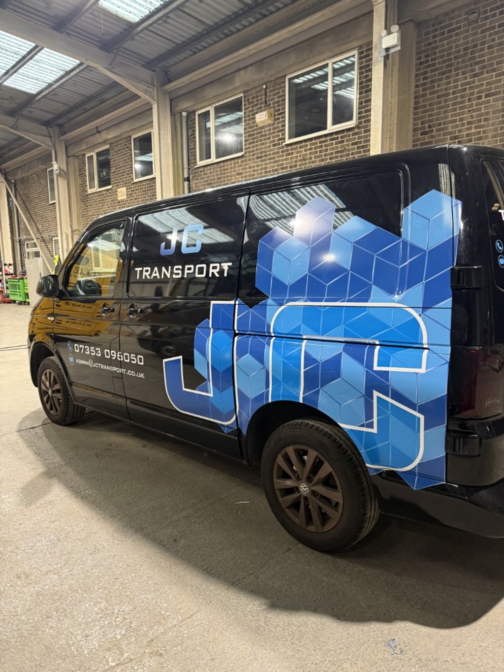 Black van with blue geometric design and branding for JC Transport, parked inside a warehouse with brick walls and high ceiling.