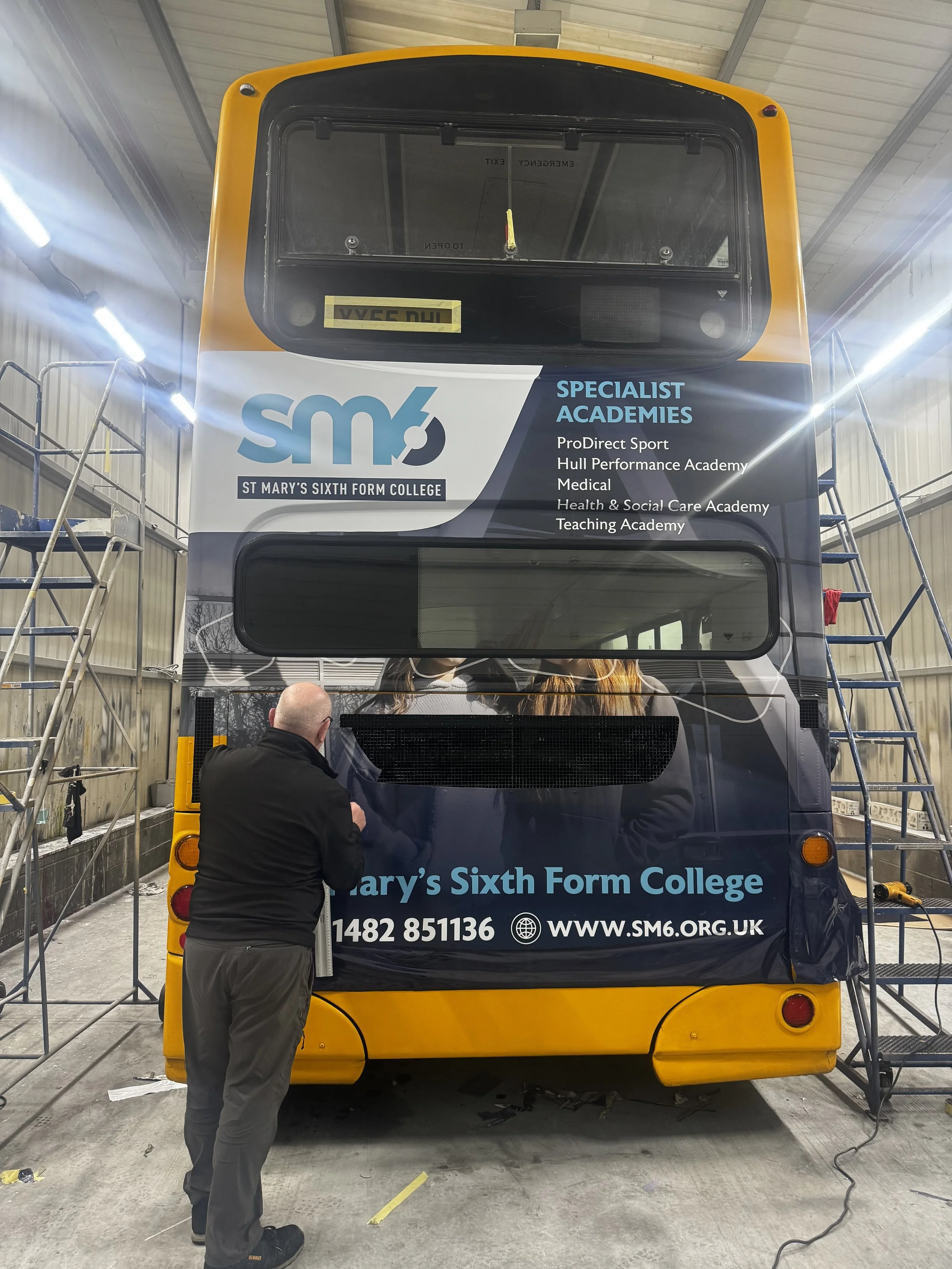 A double-decker bus with advertisements for St. Mary's Sixth Form College, featuring the college logo, contact information, and list of specialism academies. A man with a bald head wearing a black jacket stands near the rear of the bus, working on wrapping or applying decals. The bus is inside a large storage or workshop facility with scaffolding and tools around.