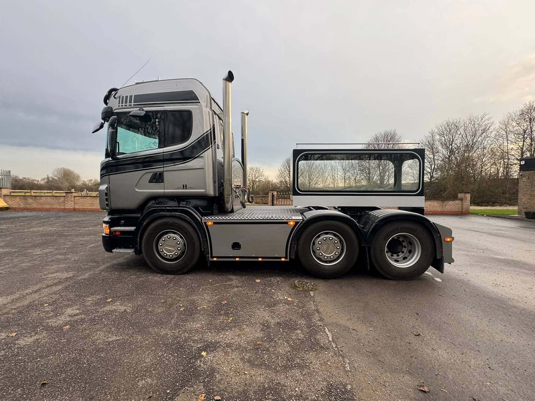 A modern, silver semi-truck tractor without a trailer attached, parked on a paved lot with a cloudy sky and leafless trees in the background.