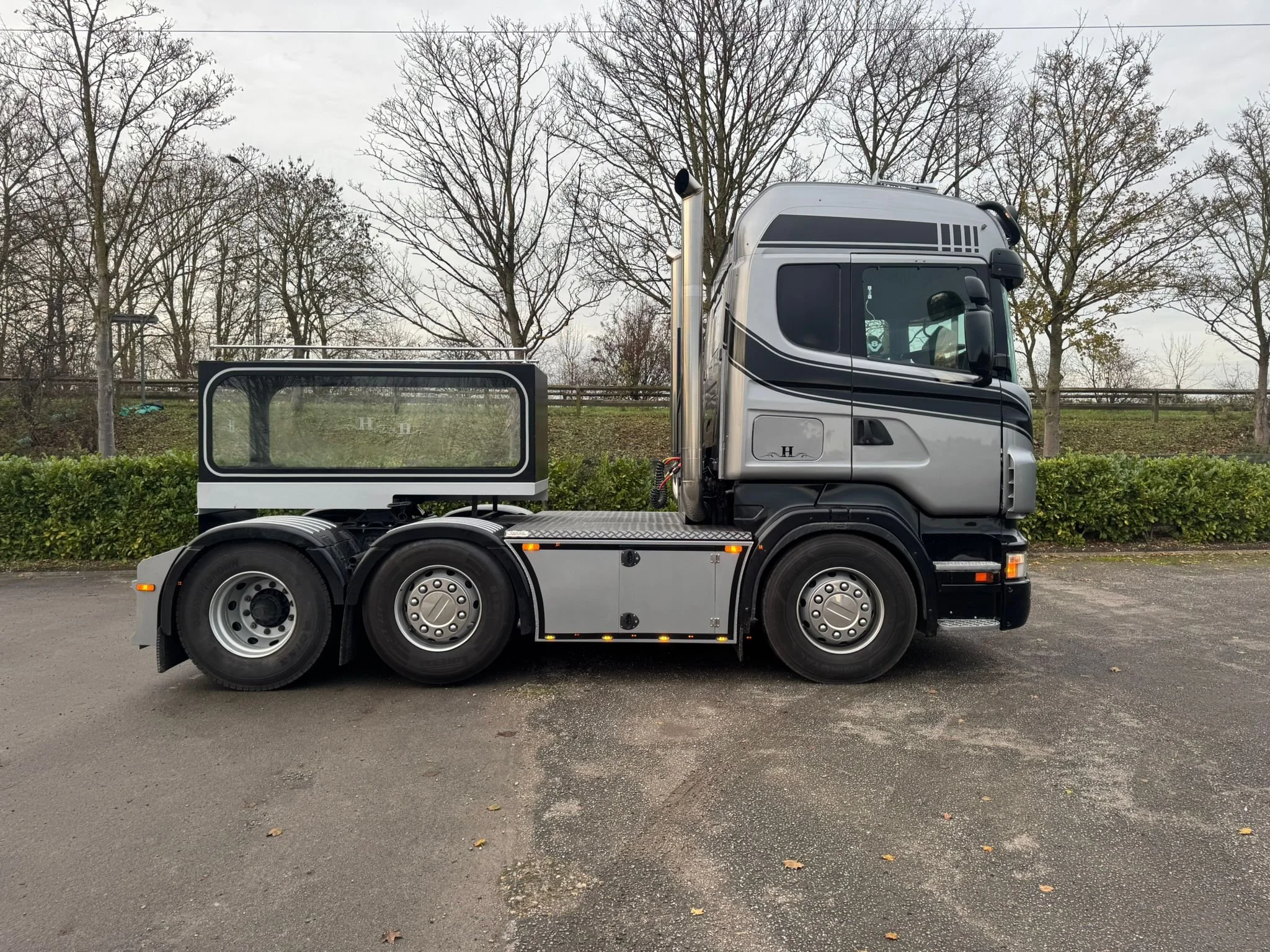 Side view of a silver semi-truck with a black stripe, parked on an asphalt lot with leafless trees and bushes in the background.
