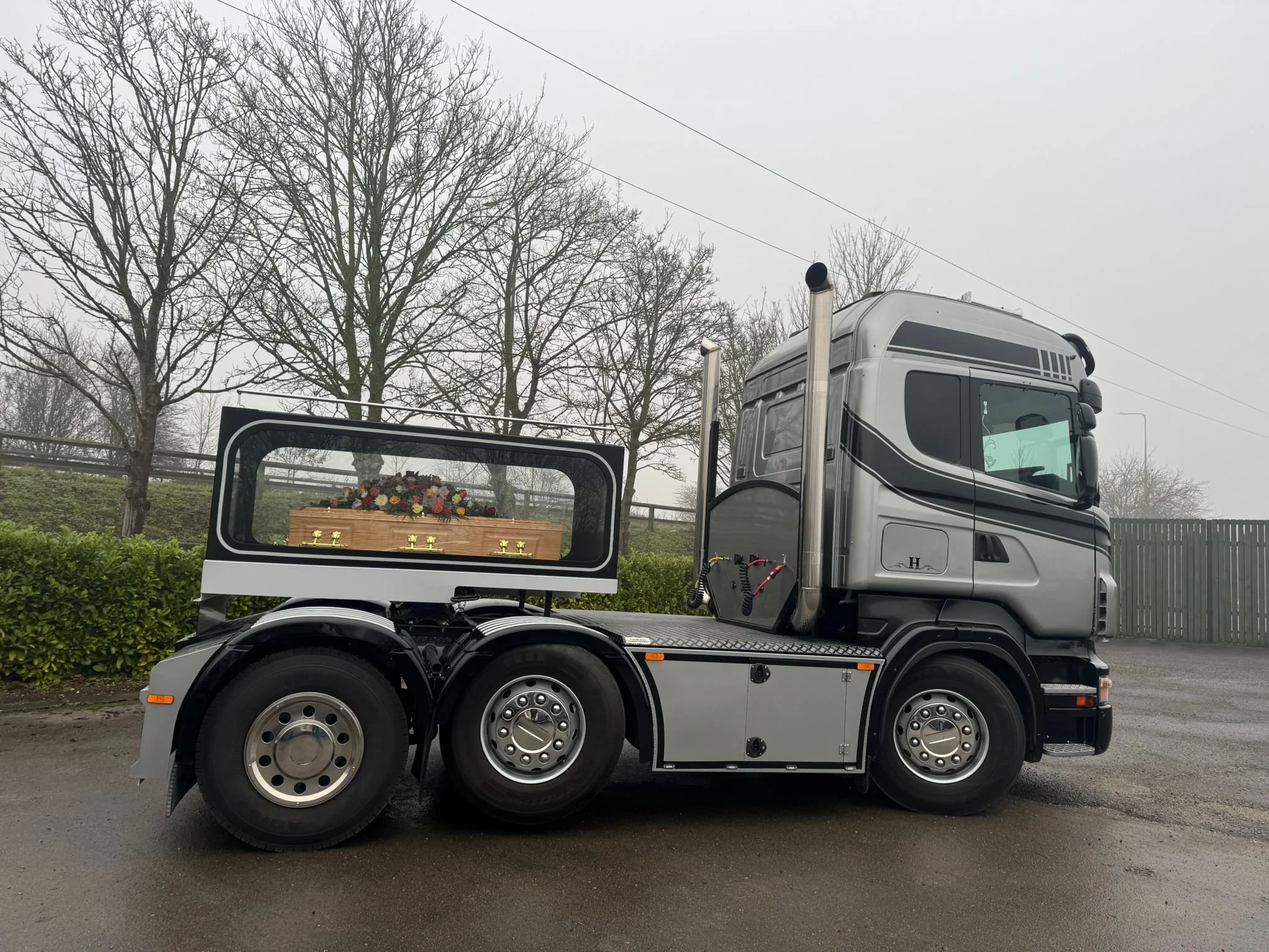 A semi-truck with a silver cab parked on a road, with a casket and flowers visible in the back compartment, alongside bare trees and overcast sky.