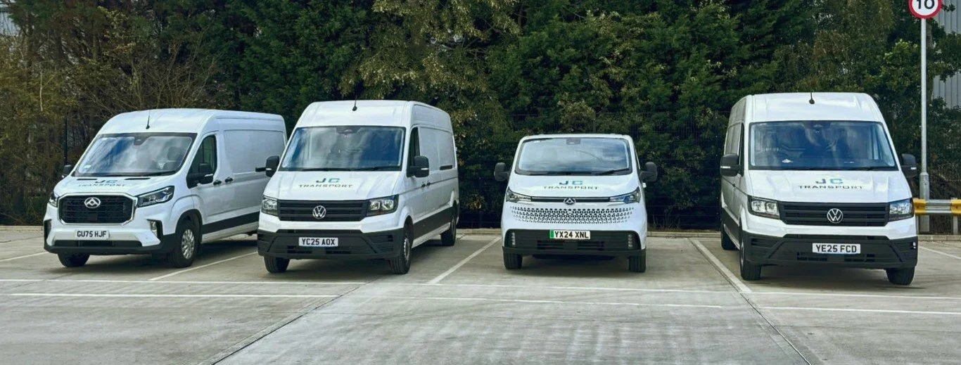Four white delivery vans parked in a lot, with green trees in the background.