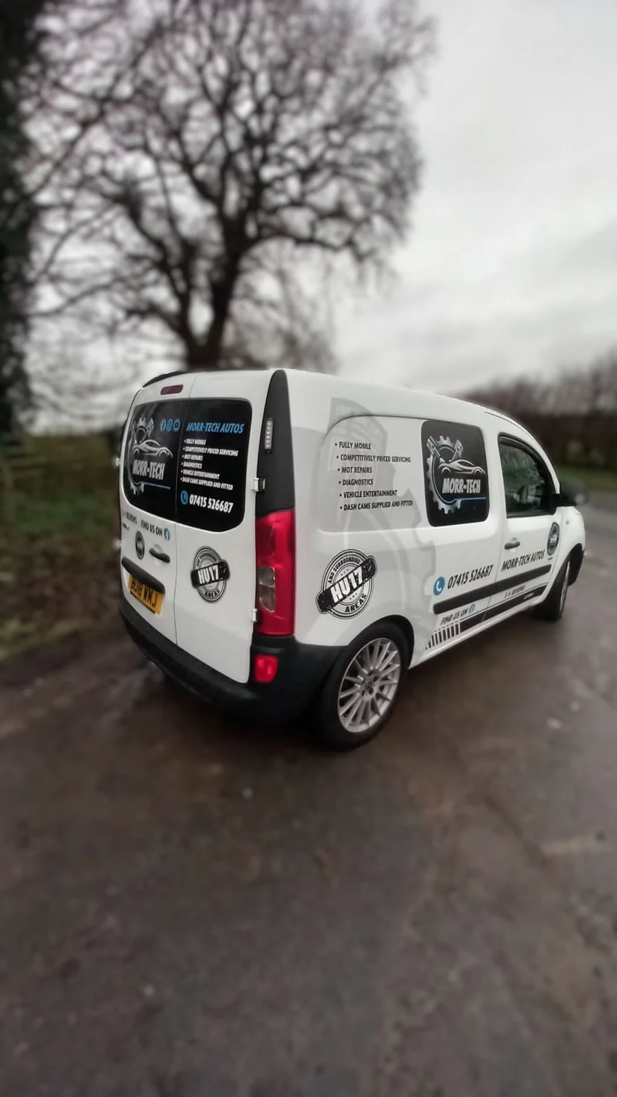 White service van with graphics and contact information parked on a dirt road near a tree with bare branches.