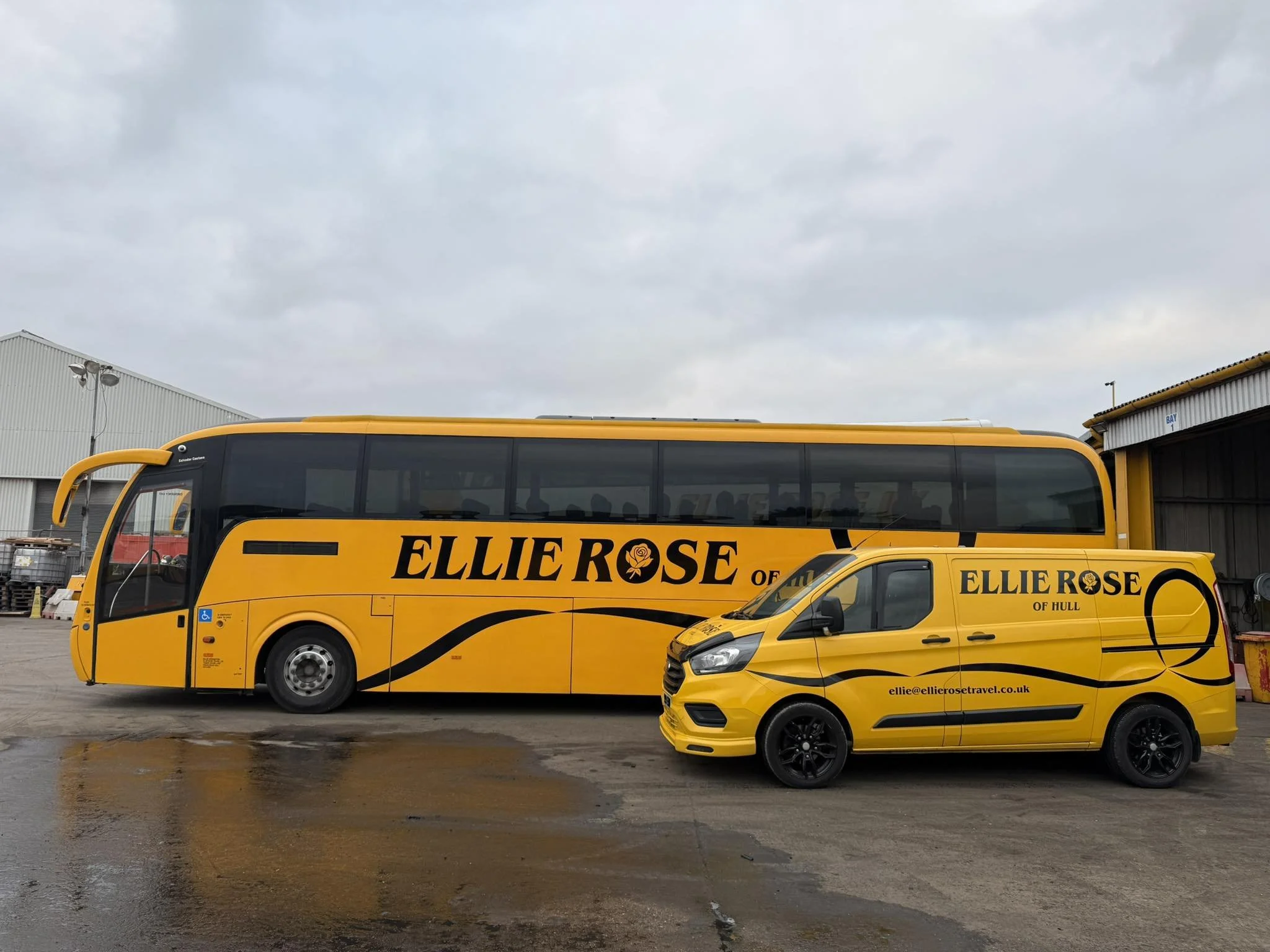 Two bright yellow vehicles, a bus and a van, parked side by side on a wet surface outside industrial buildings. Both vehicles display the word 'Ellie Rose' and branding for travel services.