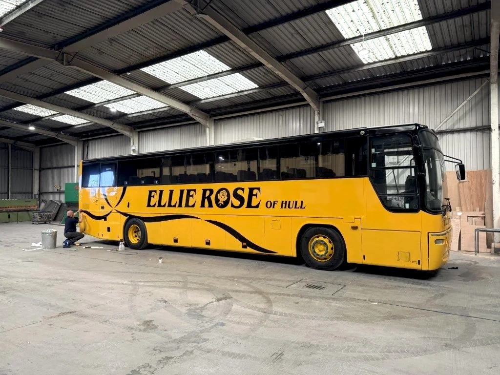 A yellow bus with black lettering that reads 'Ellie Rose of Hull' is inside a large warehouse or garage. A person is working on the bus near the back wheel, and the space has a high, metal ceiling with skylights.