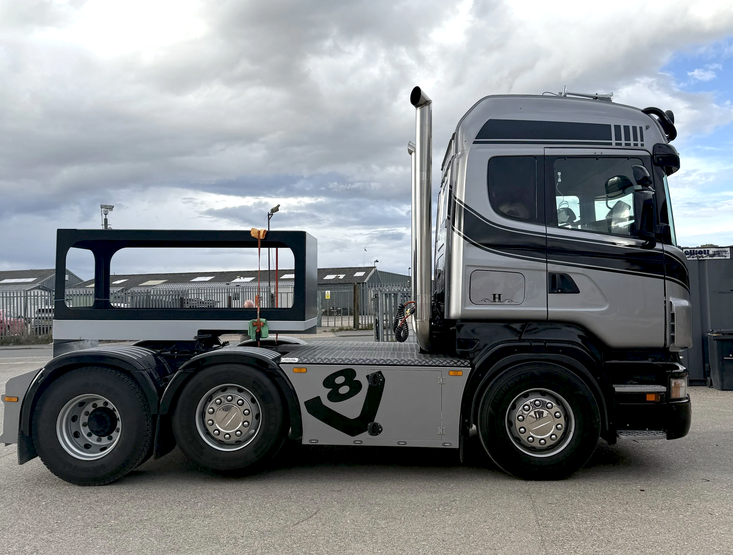 A semi-truck with an empty trailer, parked outdoors under cloudy skies, with industrial buildings in the background.