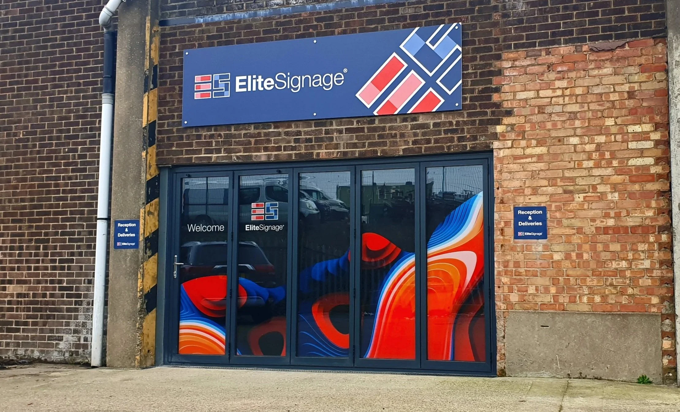 Exterior view of a brick building with signs for EliteSignage, featuring large colorful abstract graphic window display, and smaller blue signs indicating reception and deliveries.