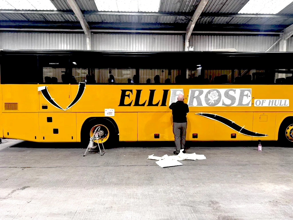 A man is painting or applying decals to a large yellow bus inside a warehouse or garage. The bus has the words 'Ellie Rose of Hull' on its side, with decorative design elements including a rose and curved lines.
