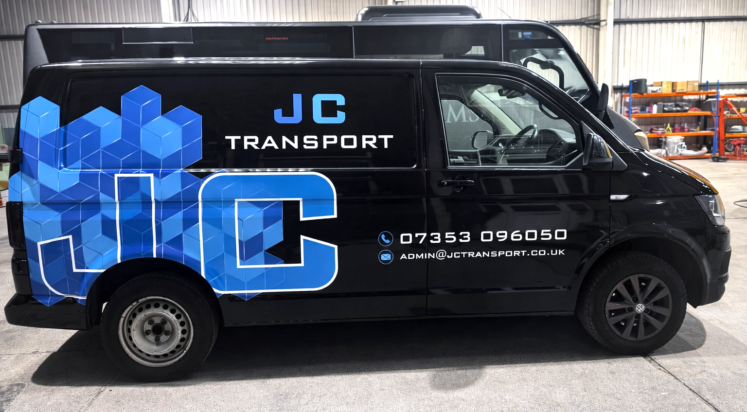Black delivery van with blue geometric graphics and JC Transport branding, parked indoors with warehouse shelving in the background.