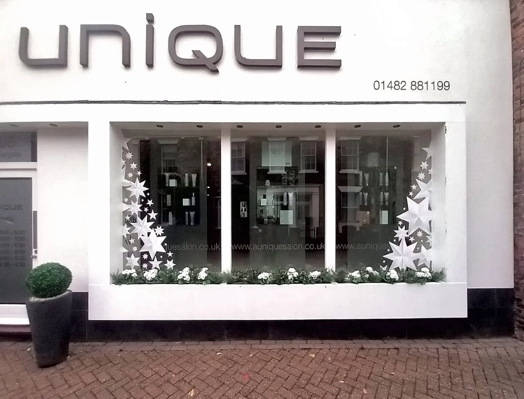 Storefront with large 'UNIQUE' sign, decorated window with white star and snowflake patterns, flower arrangement beneath window, potted bush to the left, brick sidewalk in front.