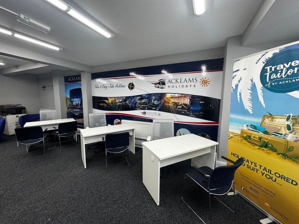 An indoor office or customer service area with tables and chairs, banners advertising Acklams Holidays and Travel Tailors, and promotional displays for travel vacation packages.