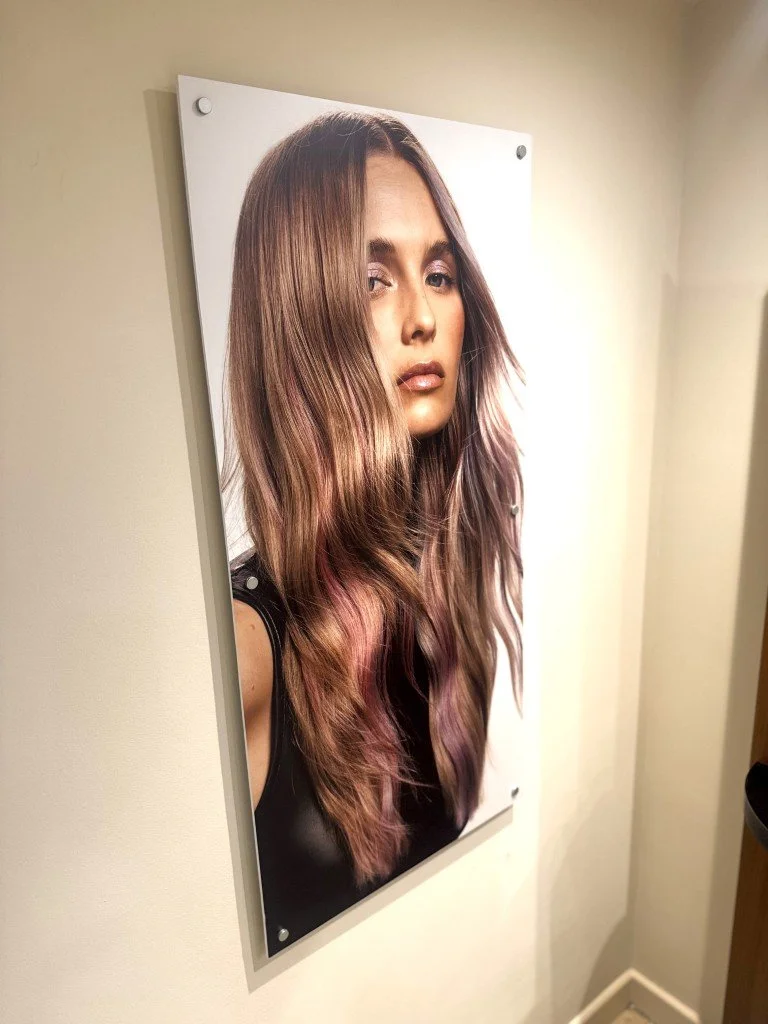 Photograph of a woman with long wavy hair featuring brunette and pink highlights, wearing a black sleeveless top, displayed on a wall.