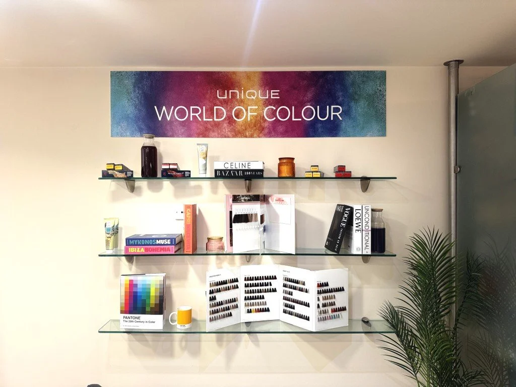 Decorative wall display with three glass shelves holding nail polish samples, books, a Pantone color guide, a mug, small decorative jars, a color swatch, and a colorful sign that says 'UNIQUE WORLD OF COLOUR'. A potted plant is on the right side.