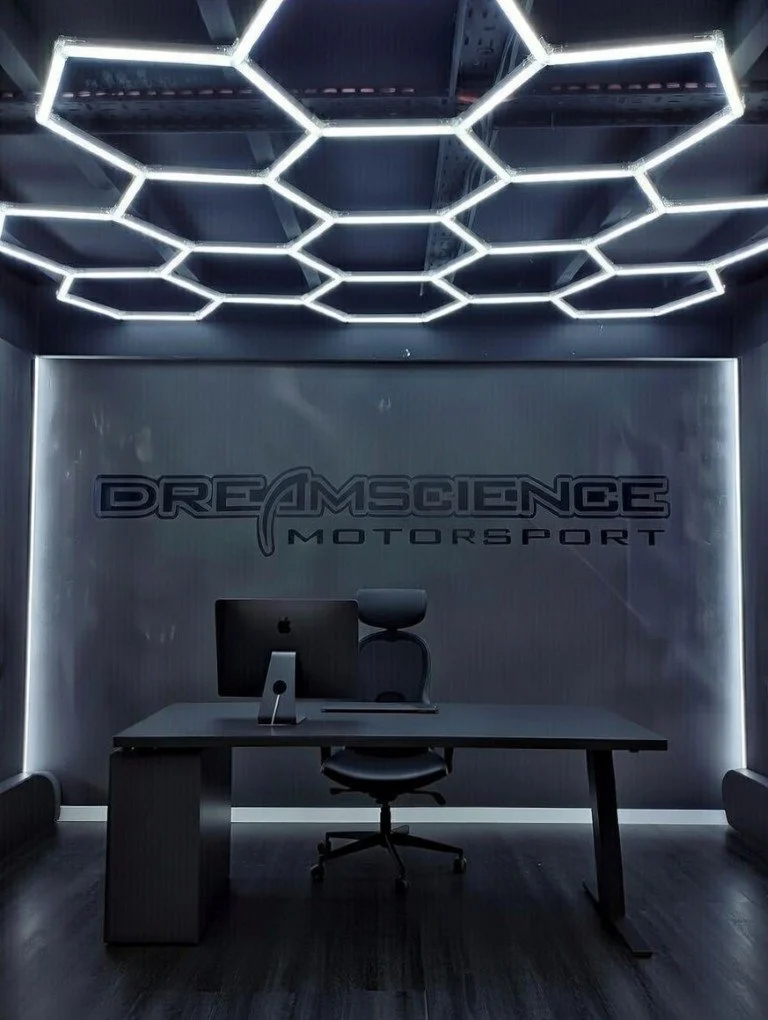 Modern office with a minimalist desk, black office chair, iMac computer, and illuminated geometric hexagon ceiling design in a dark room with a logo on the wall reading 'Dream Science Motorsport'.