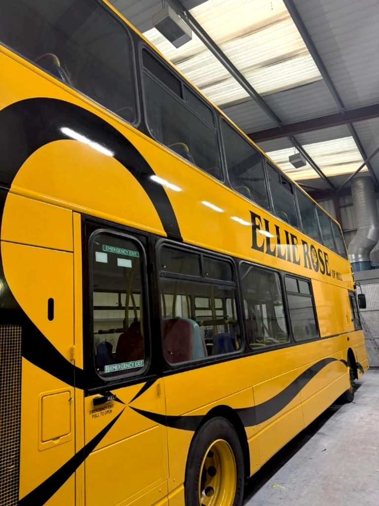 Bright yellow double-decker bus with black lettering reading "Elisabeth Rose of Mill" parked inside a warehouse with metal roof and large vents.