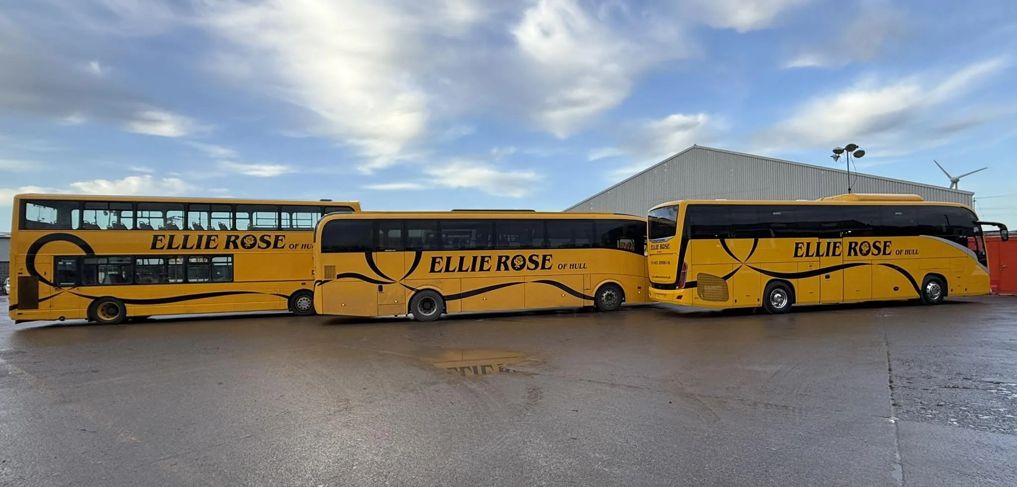 Three yellow double-decker buses with black lettering and designs, parked outdoors on a wet pavement with a cloudy sky and a large industrial building in the background.