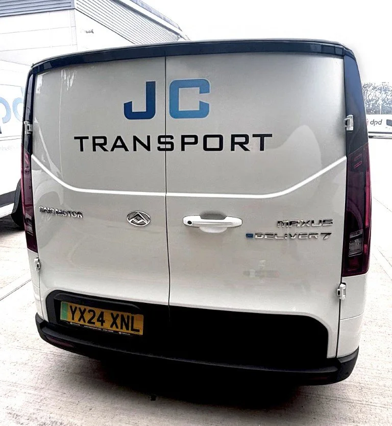 Rear view of a white JC Transport delivery van with a UK license plate, parked indoors. The back of the van displays the JC Transport logo, and logos for Maxus Deliver 7 and Sadro Motor.