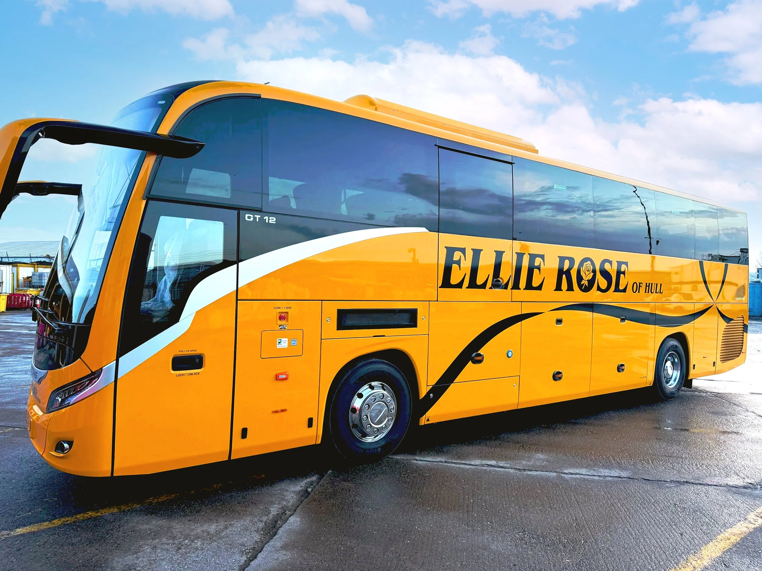 Yellow tour bus with the name "Ellie Rose of Hull" on its side, parked on a wet pavement on a partly cloudy day.