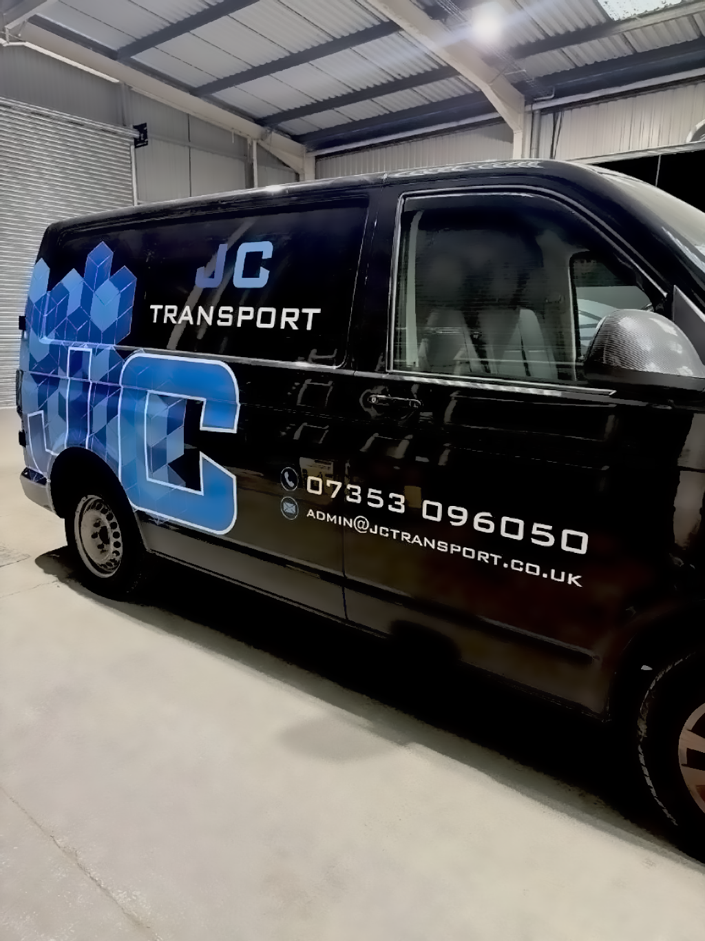 Black delivery van with blue and white graphics and text, parked inside a warehouse, advertising JC Transport with contact details.