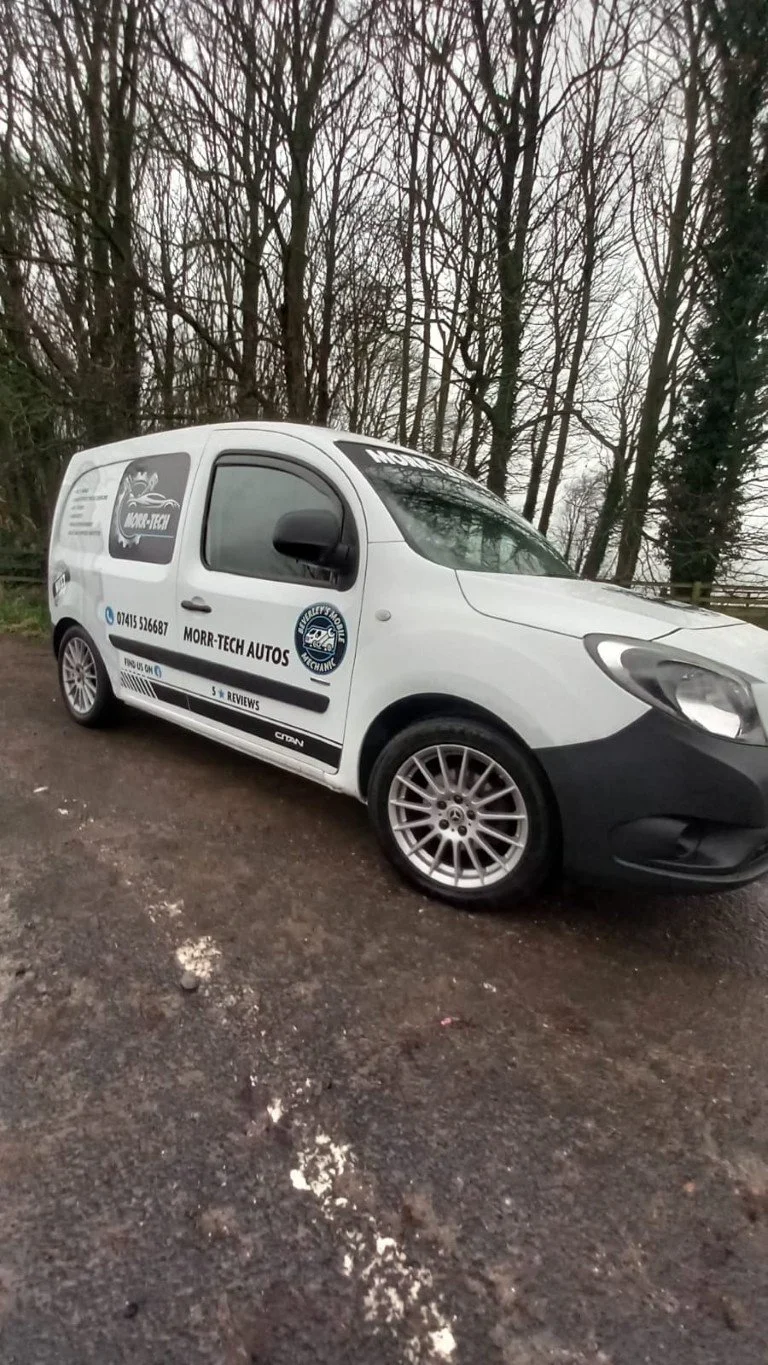 White commercial van with black and white branding for Morr-Tech Autos parked on a dirt surface, surrounded by leafless trees.