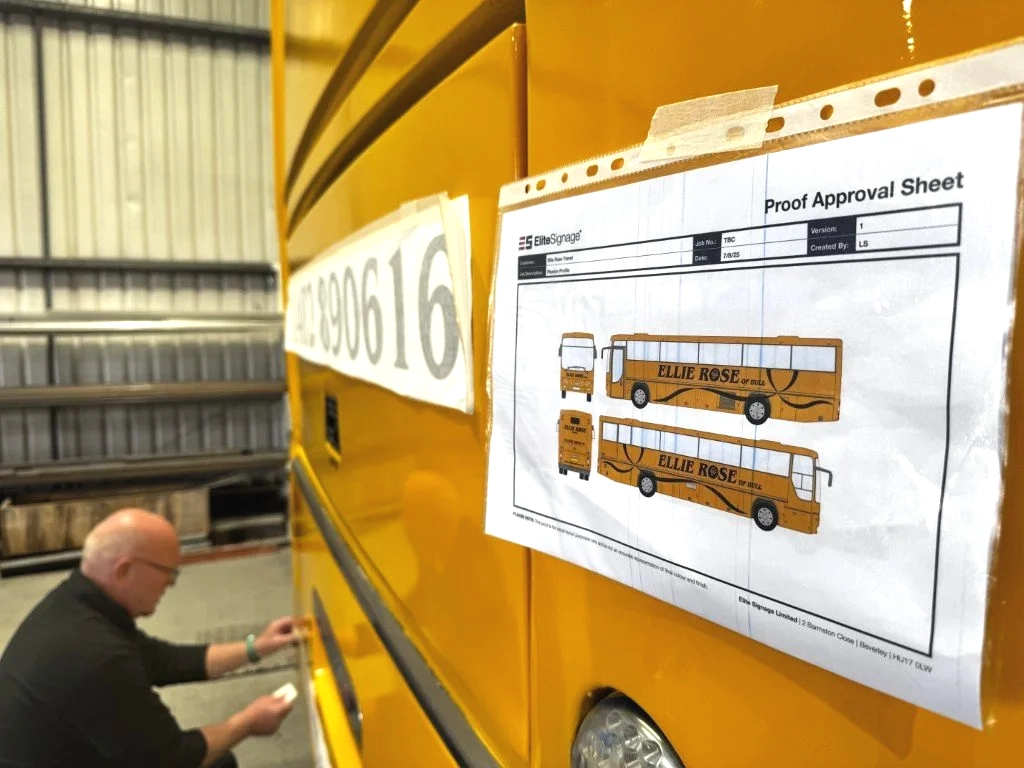 A man in black shirt working on a yellow bus inside a warehouse, with a proof approval sheet attached to the bus showing the design of the bus labeled "Ellie Rose."