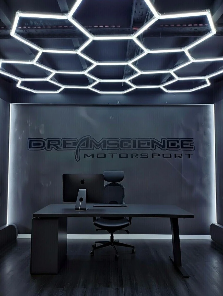 Modern office with a black desk, black ergonomic chair, computer monitor, and a large wall sign that reads "Dreamscience Motorsport". Hexagonal LED light fixtures are on the ceiling.