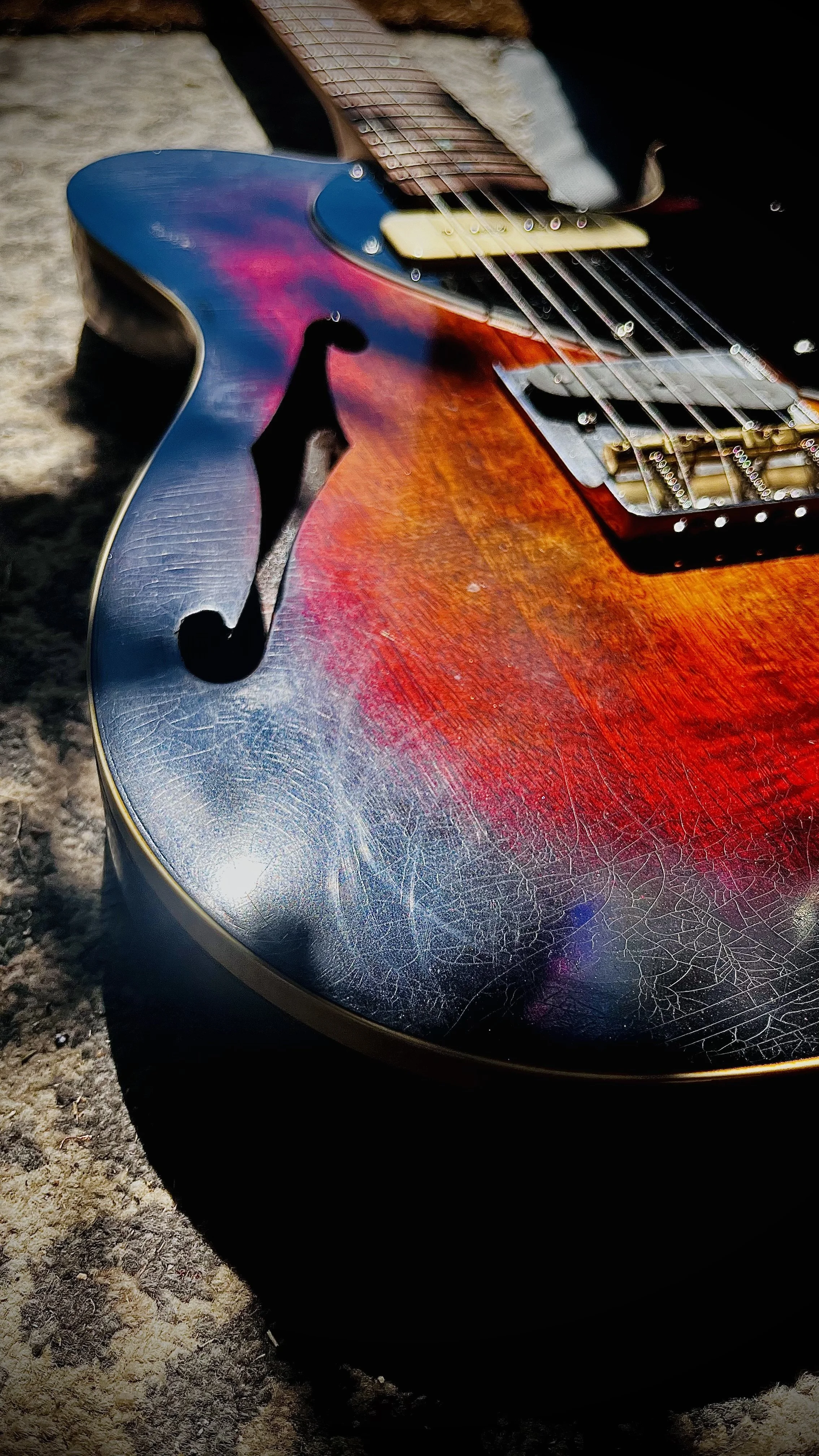 Close-up photograph of a vintage sunburst electric guitar with a crackled finish, lying on a textured surface, showing the body, strings, and f-holes.