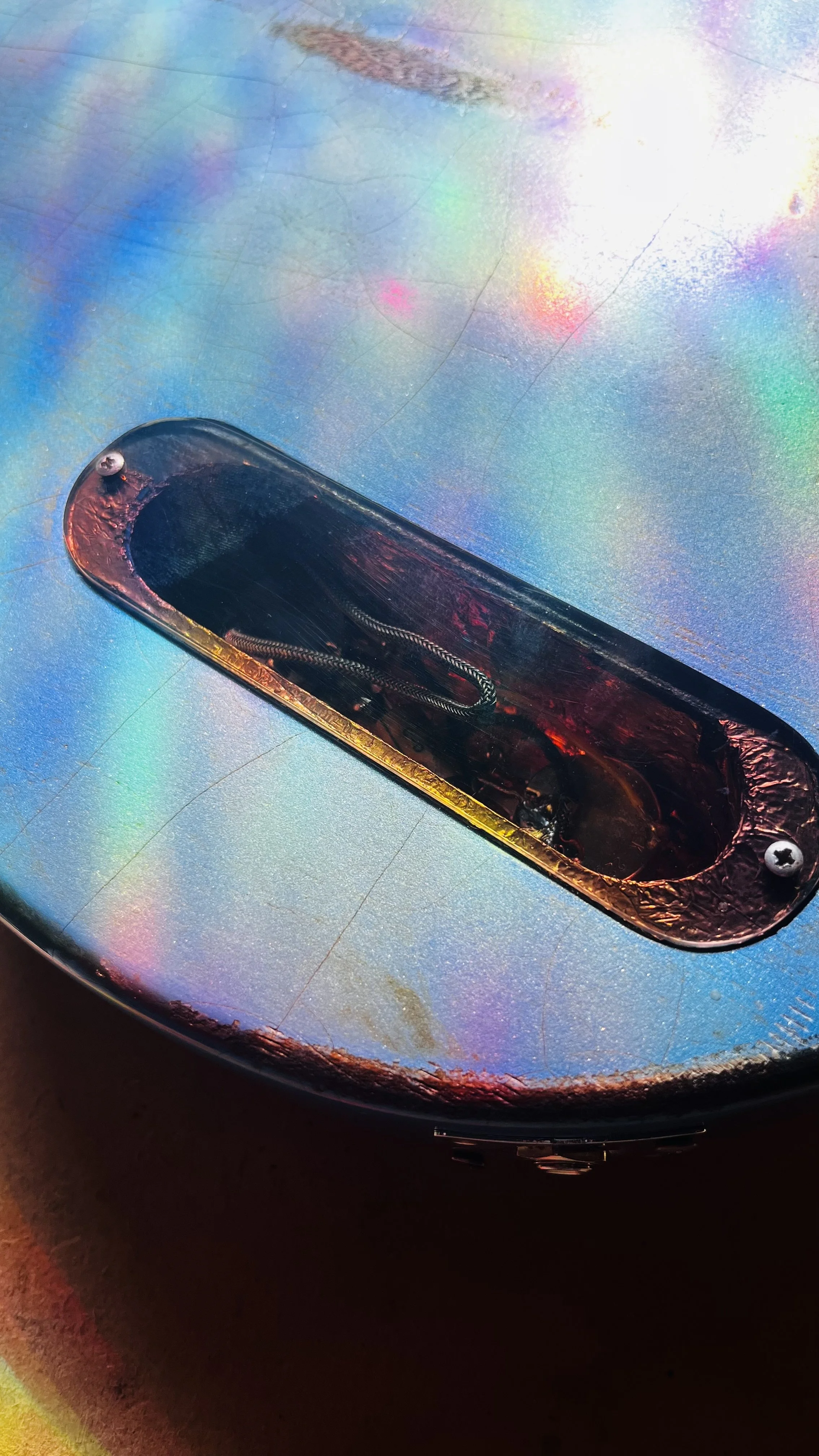 Close-up of a metallic, iridescent surface with a recessed area containing a black component with a visible zipper, screws, and a wire inside.