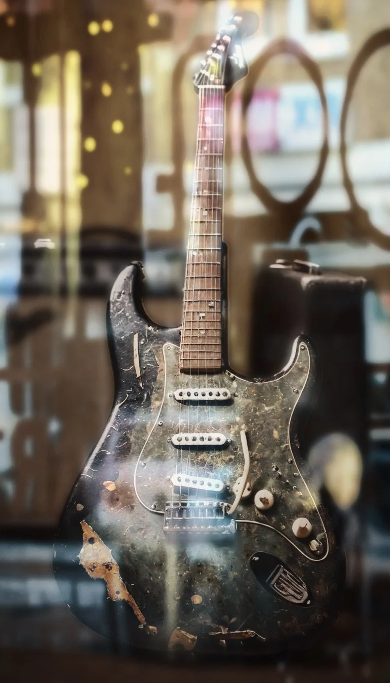 An old, worn electric guitar with a black finish, multiple scratches, and peeling paint, displayed inside a glass case.