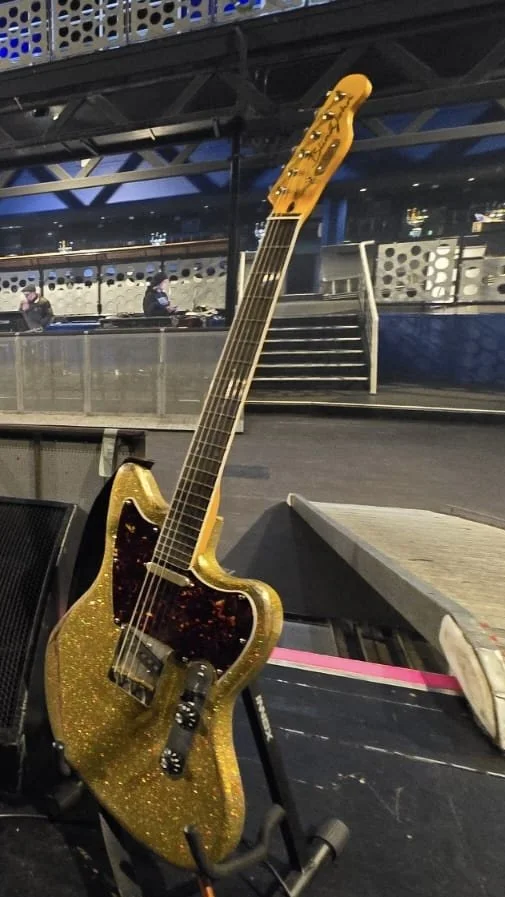 Electric bass guitar with a gold glitter finish and black pickguard resting on a stand next to a keyboard in an indoor setting at night.