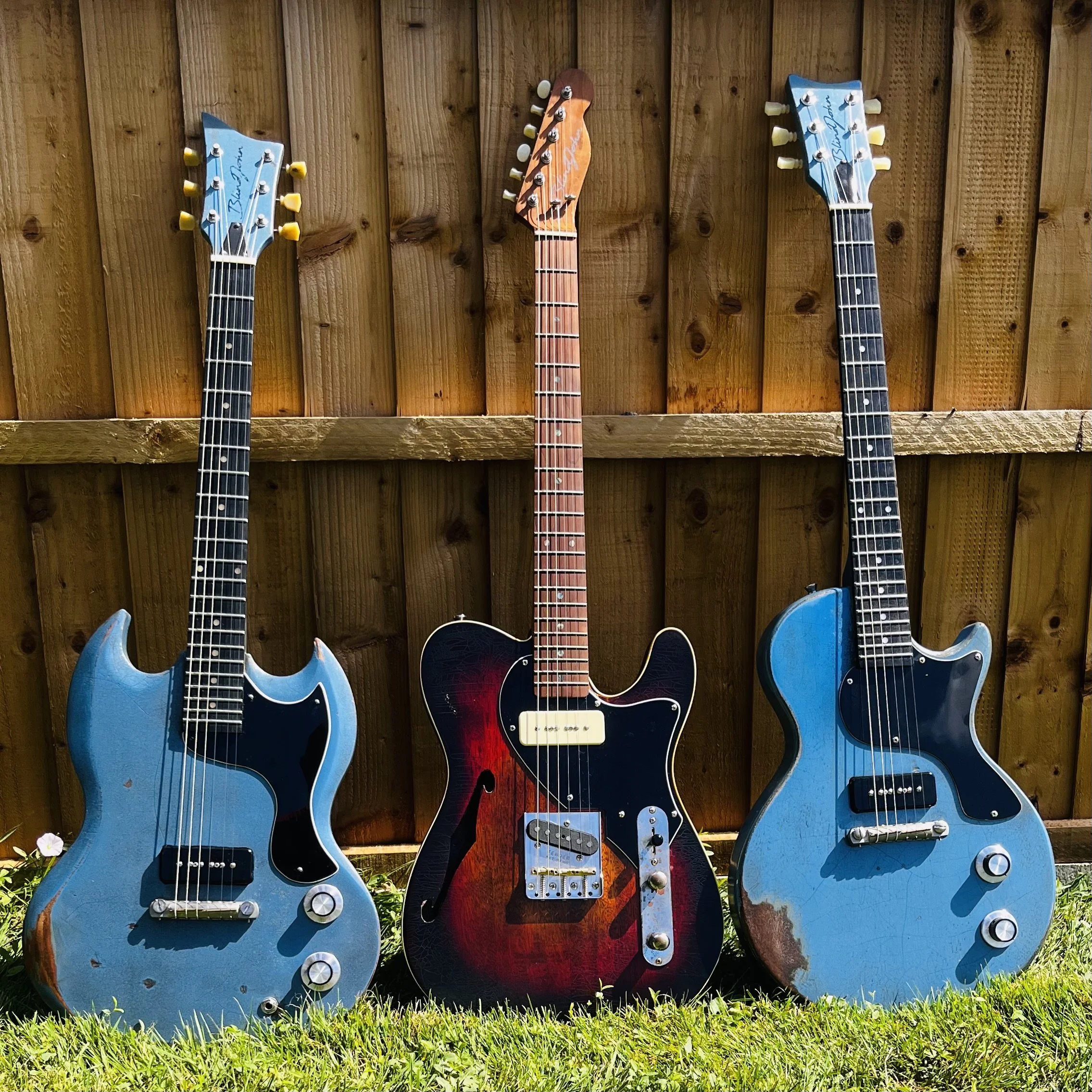 Three vintage electric guitars, two blue and one sunburst, are leaning against a wooden fence on a grassy lawn.