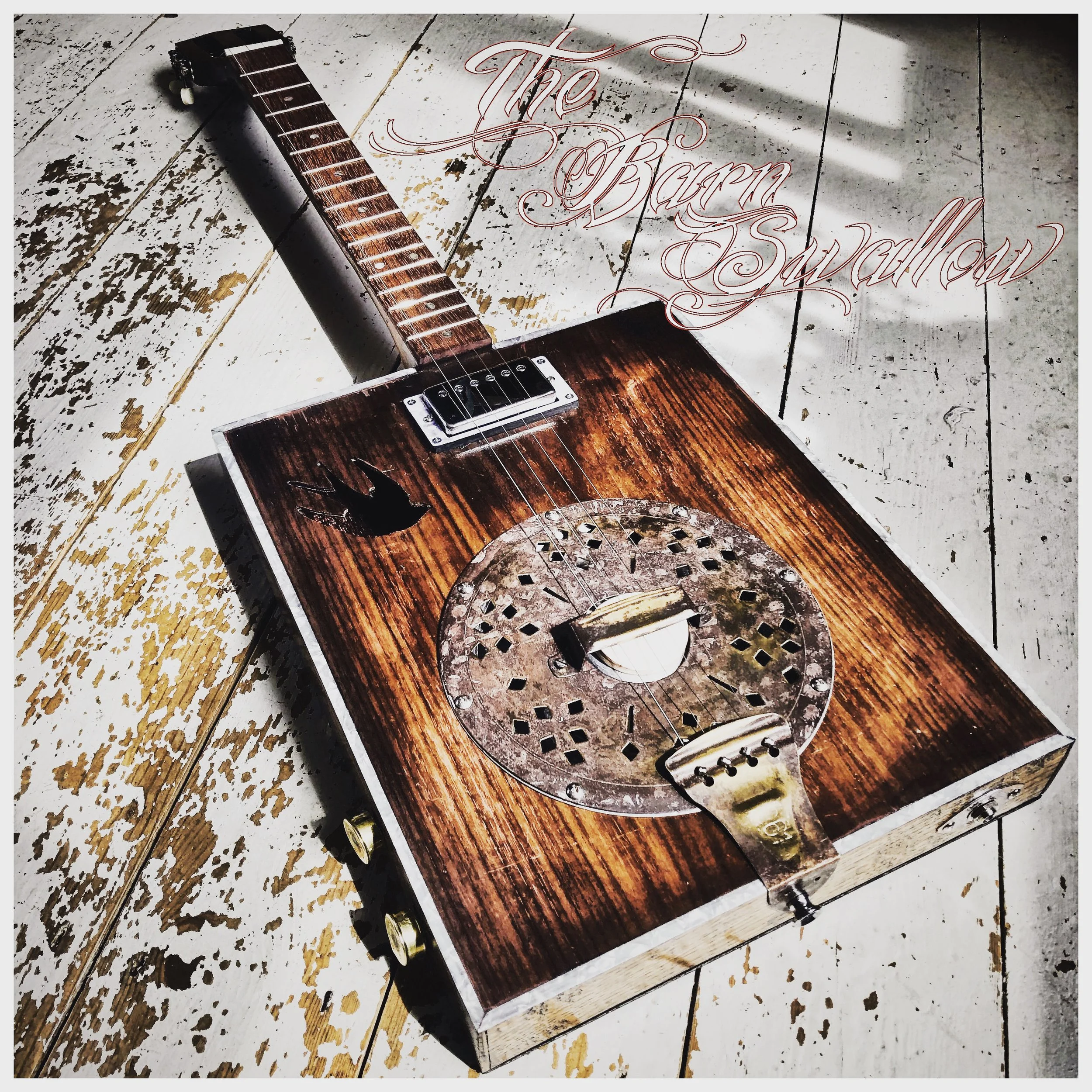 A wooden cigar-box guitar with metal tuning pegs and a pickup, resting on a distressed white wooden surface, with the text 'The Barn Swallow' in decorative cursive font.