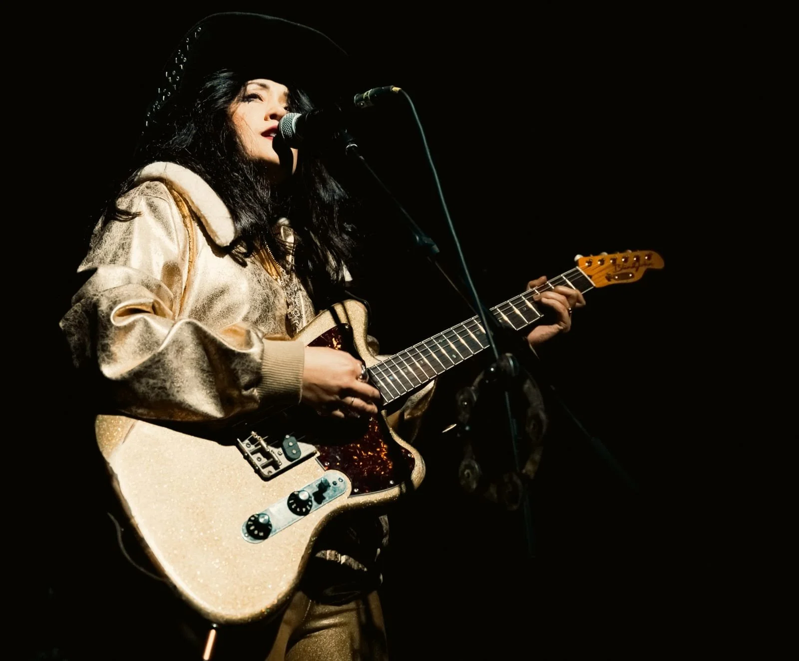 A woman with long black hair wearing a large black hat and gold jacket playing an electric guitar and singing into a microphone on a dark stage.