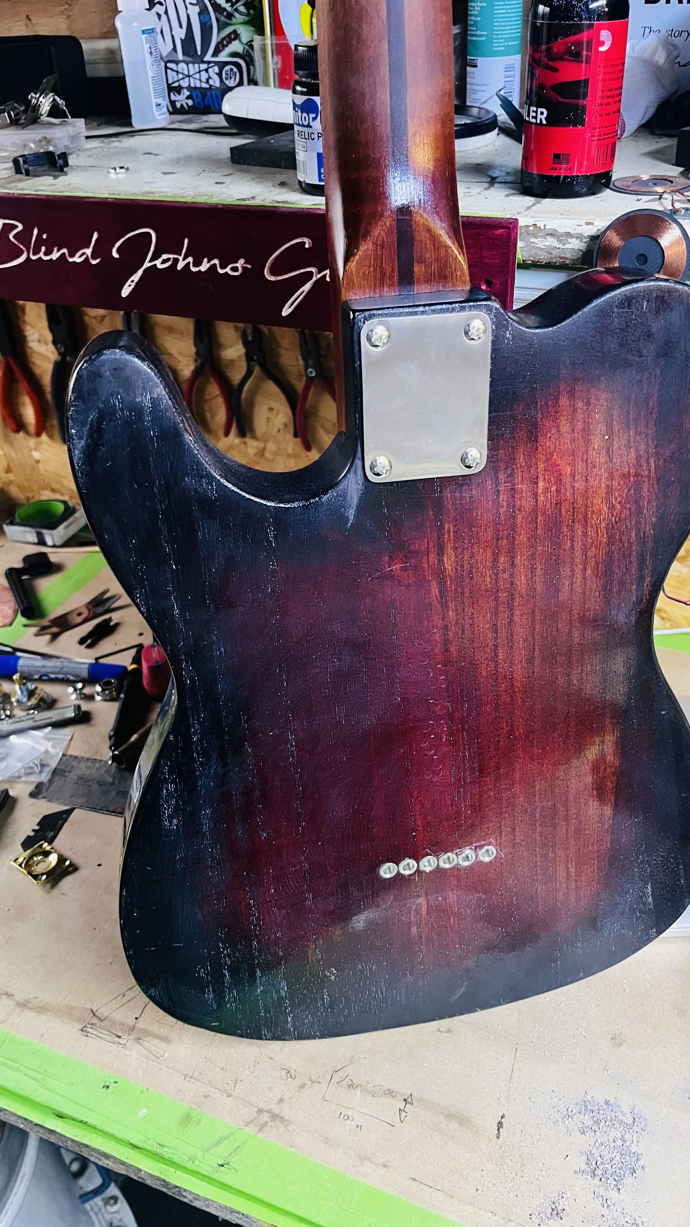 Close-up of the back of a red and black electric guitar in a workshop, showing the neck joint secured with four screws and a string of six tuning pegs on the body.
