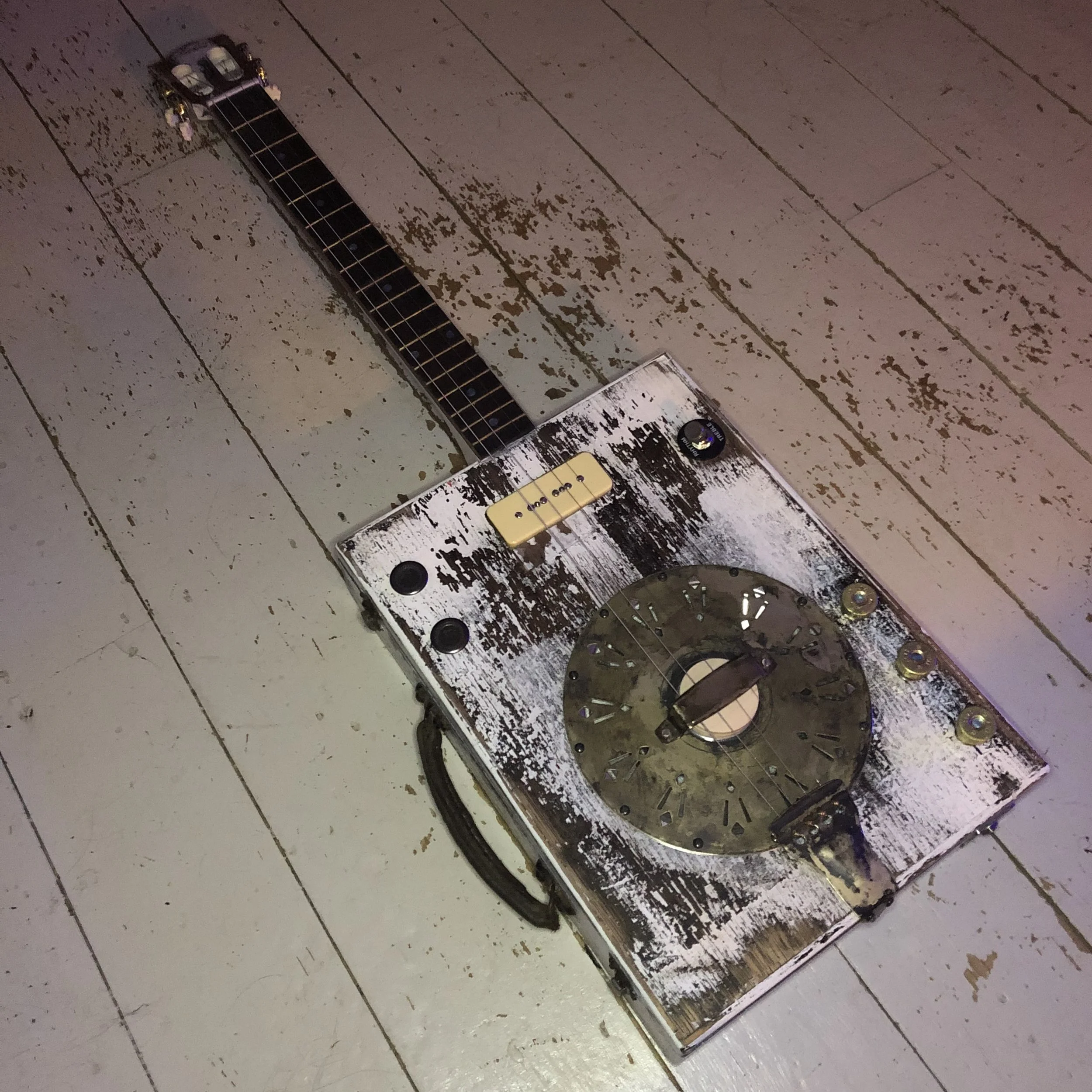 A homemade guitar with a vintage, distressed look, featuring a rectangular body with a circular metal plate and tuning components, placed on a white, weathered wooden floor.