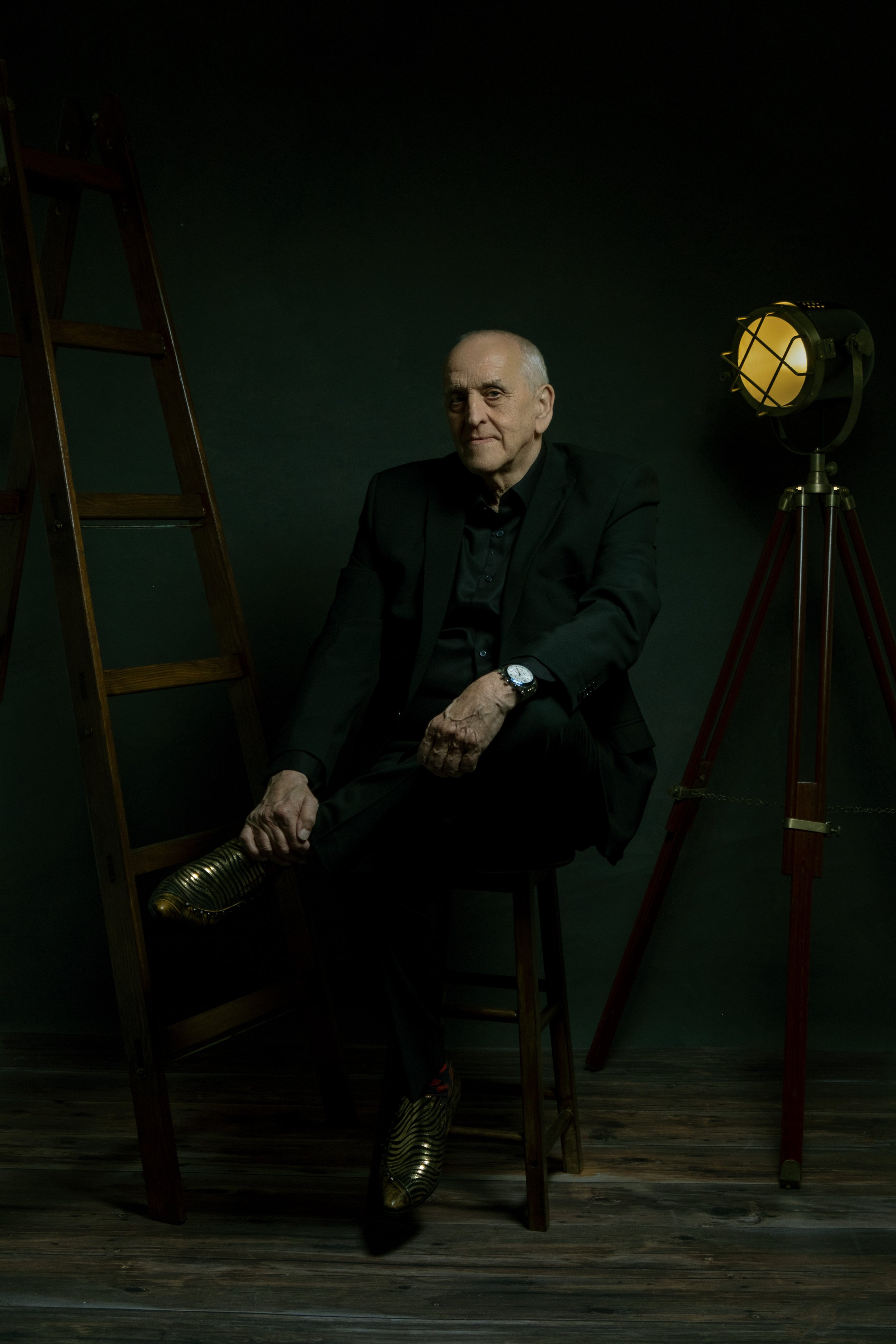 Graham Short Portrait Photo. Graham sitting on a chair in a dark green room, next to a ladder, like in a studio.