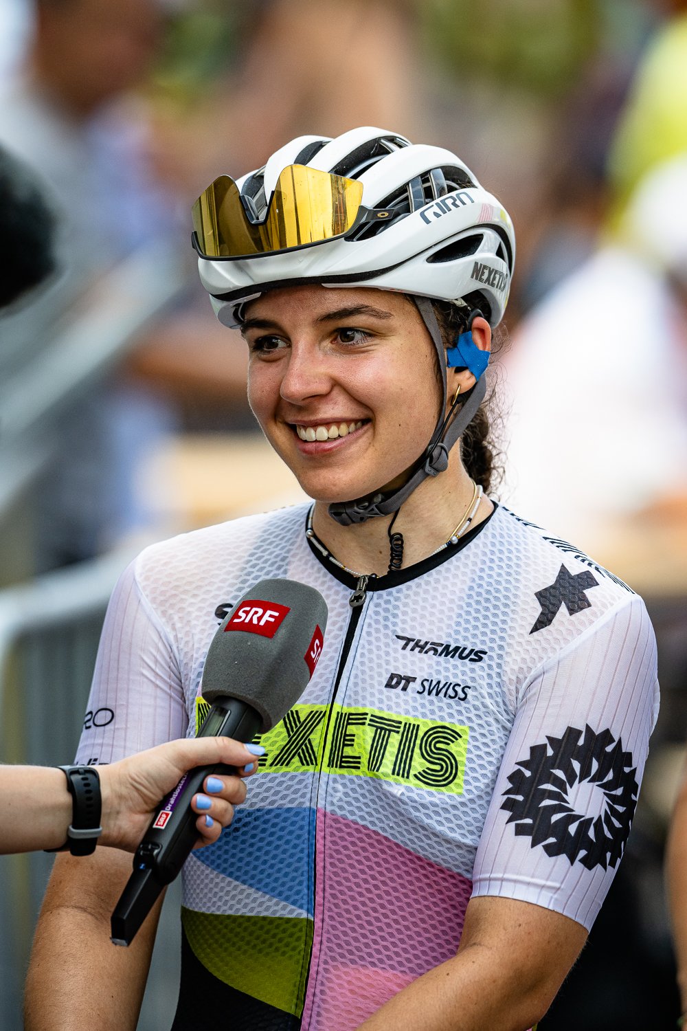 Junge Radfahrerin im Fahrradhelm wird interviewt, im Hintergrund Zuschauer, trägt ein Teamtrikot mit verschiedenen Sponsoren.