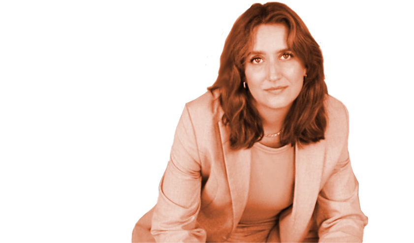 Eine Frau mit braunen, gewellten Haaren trägt eine beige Jacke und sitzt nach vorne gerichtet.