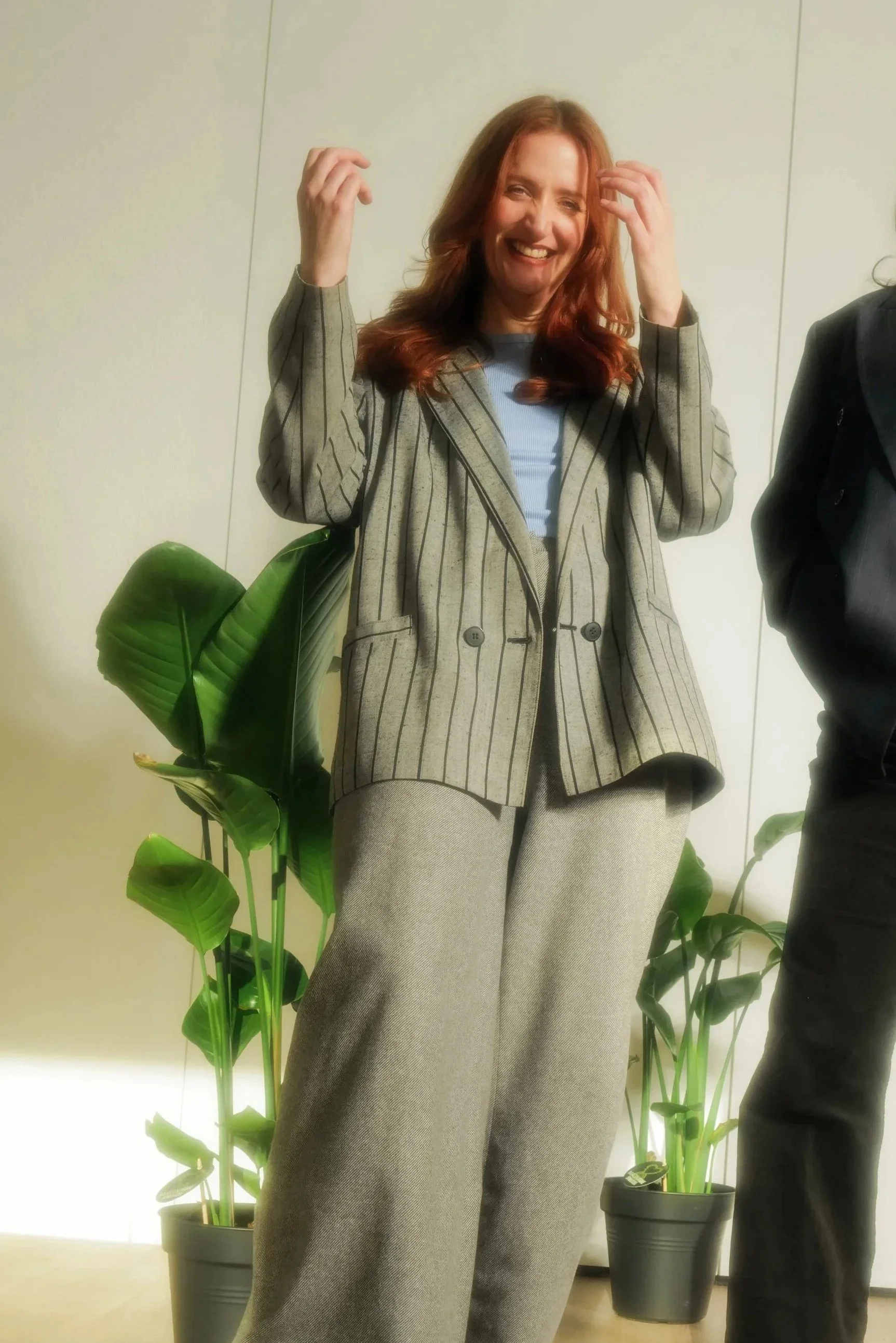 Lächelnde Frau mit roten Haaren, trägt einen grauen, gestreiften Blazer und passende Hosen, steht neben einer Pflanze in einem Topf, im Hintergrund eine weiße Wand.