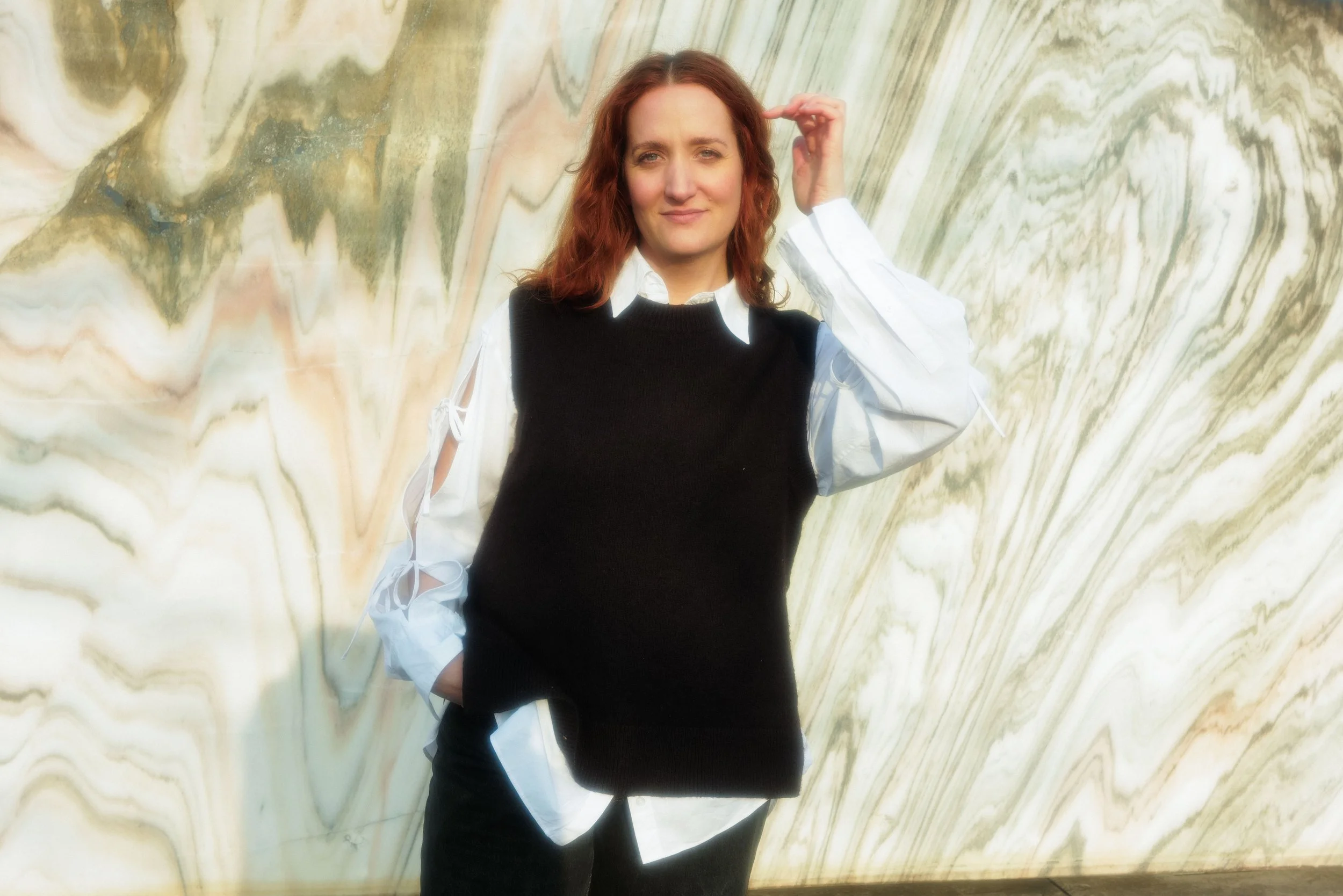 Eine Frau mit roten Haaren steht vor einer Marmorwand und trägt einen schwarzen Pullover über einem weißen Hemd.