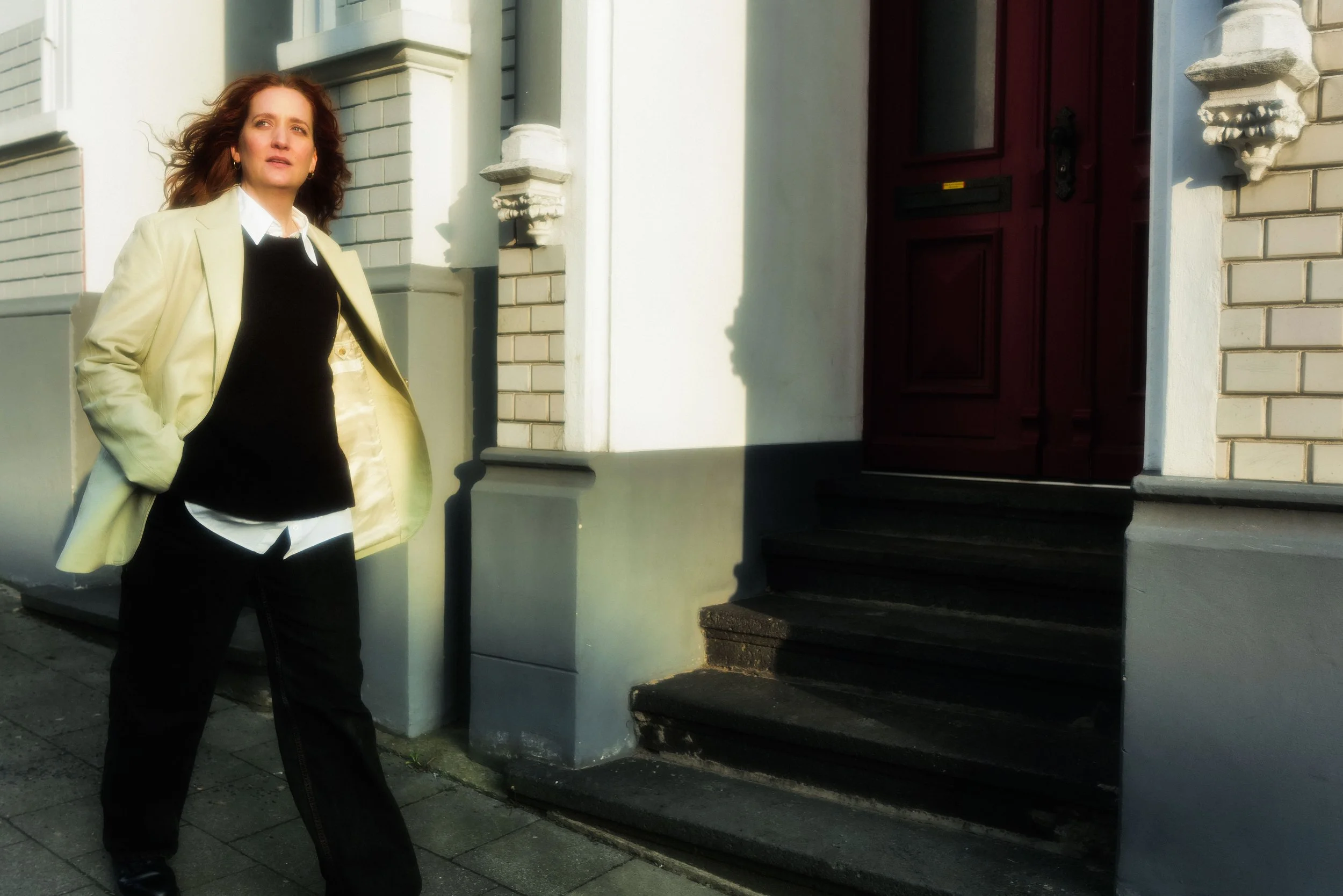 Eine Frau mit braunen, lockigen Haaren trägt eine schwarze Hose und eine schwarze Bluse, eine helle Jacke liegt offen auf ihren Schultern, sie steht vor einem Haus mit roten Türen und scheinbar in moderner Kleidung.