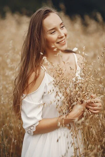 A young woman with long brown hair smiling and holding a bouquet of dried wildflowers in a field of tall, dry grass.
