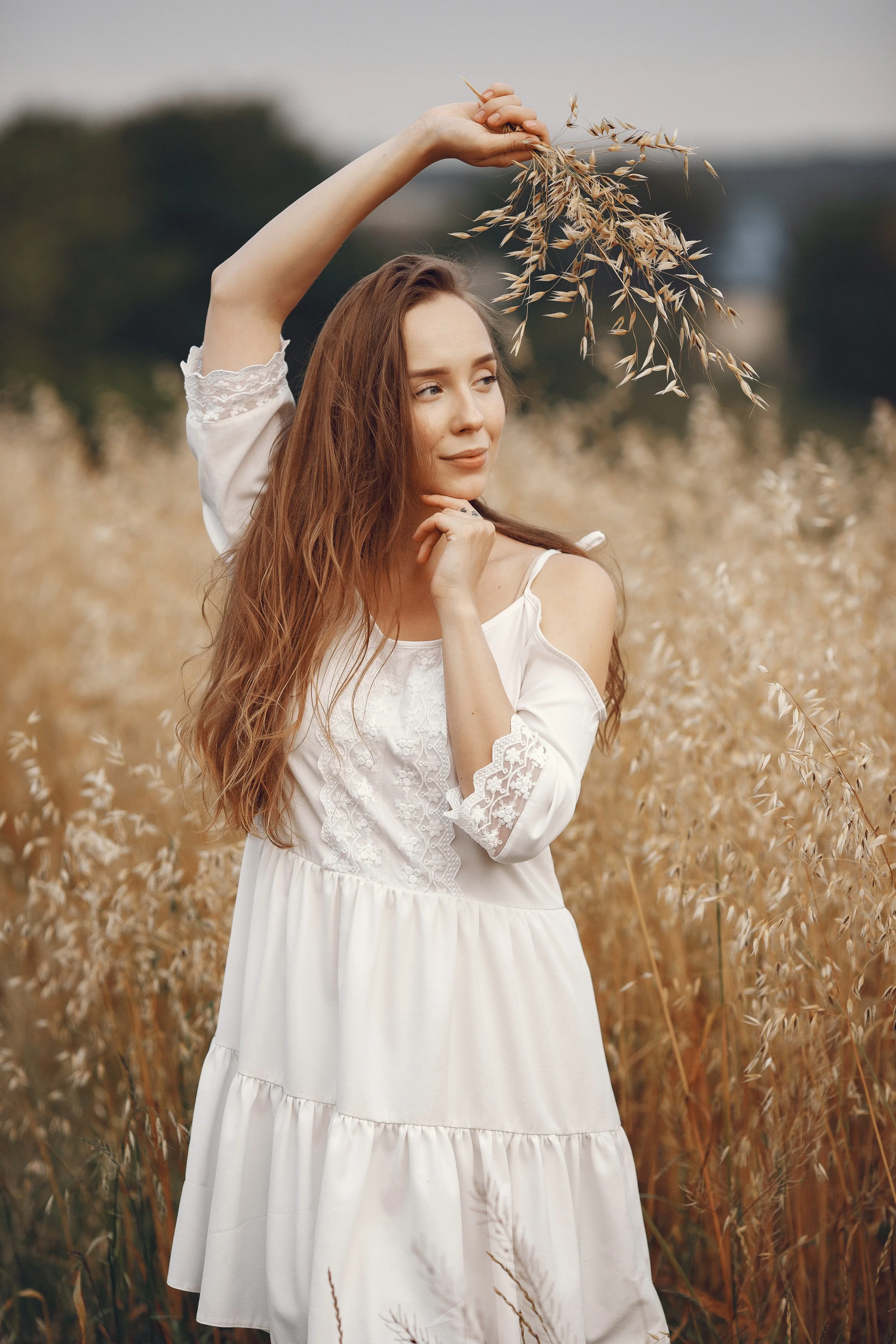 A woman with long, wavy auburn hair wearing a white dress with lace detail, standing in a field of tall, golden grass, holding a stalk of grass above her head with a serene expression.