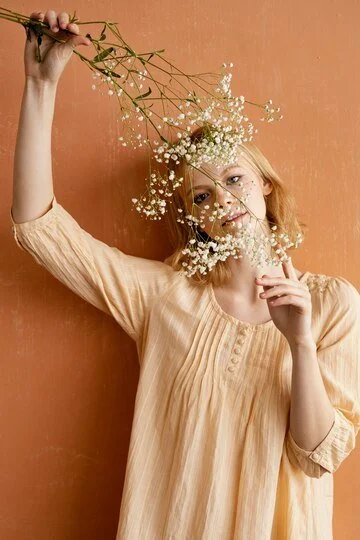 A young woman with blonde hair holds a branch of white baby's breath flowers above her head while standing against an orange wall.