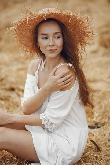 A young woman sitting outdoors on a dry, grassy field, wearing a wide-brimmed straw hat and a white dress with lace details on the sleeves, looking to the side with a gentle expression.