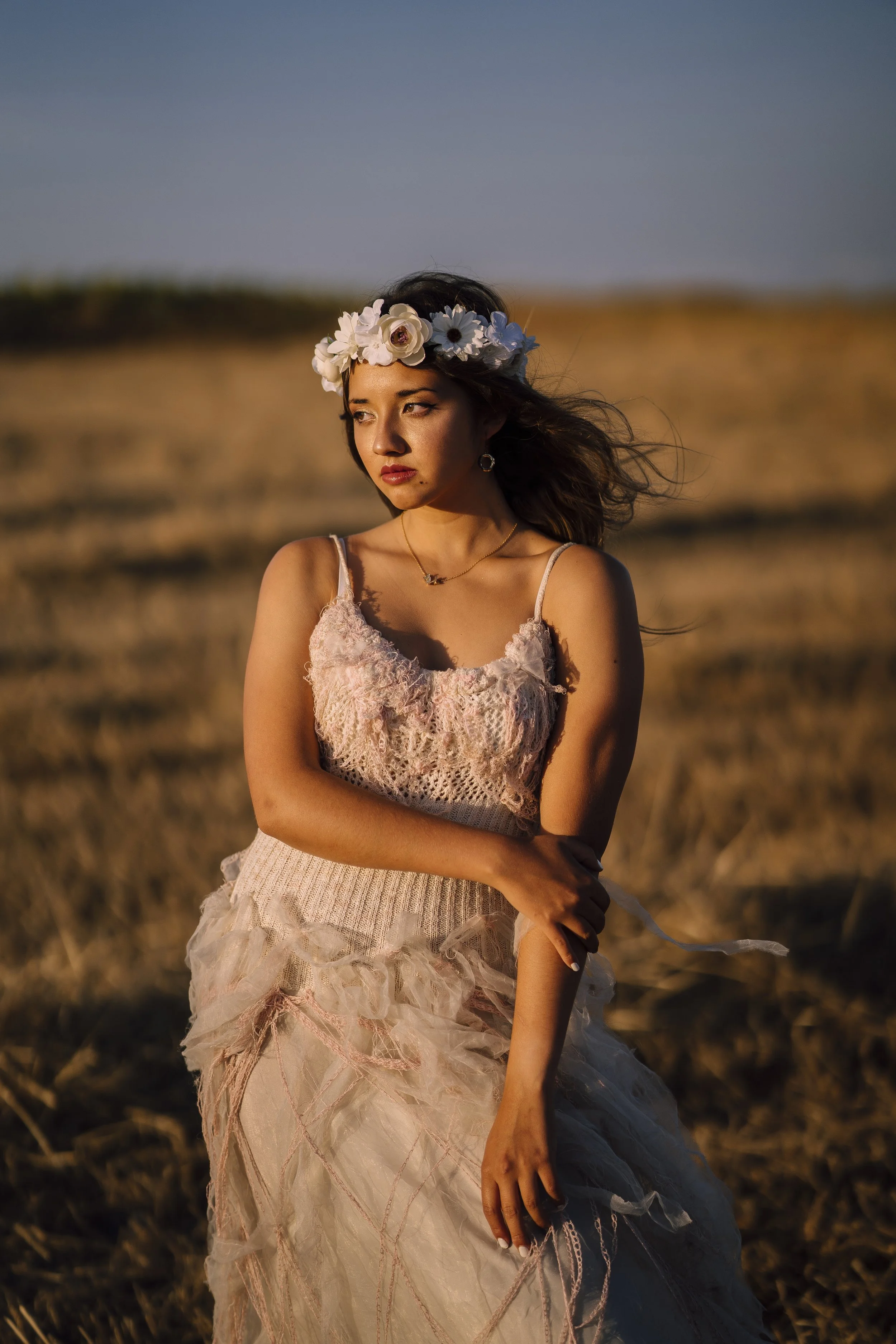 A woman in a cream-colored, lacy dress standing in a field with dried grass during sunset, wearing a flower crown and looking into the distance.