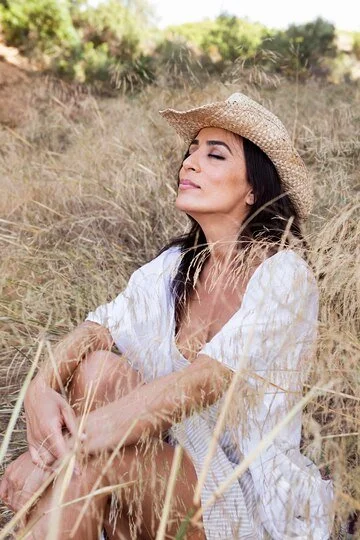 Woman wearing a straw hat sitting in a field of tall grass with a calm expression and closed eyes.