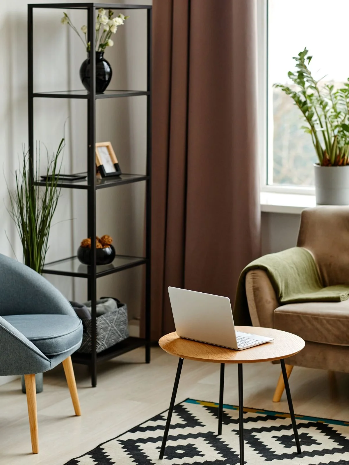 Living room with a laptop on a small wooden table, a beige armchair with a green throw blanket, a gray chair, a black metal shelf with decorative vases and picture frames, a window with brown curtains, and potted plants.