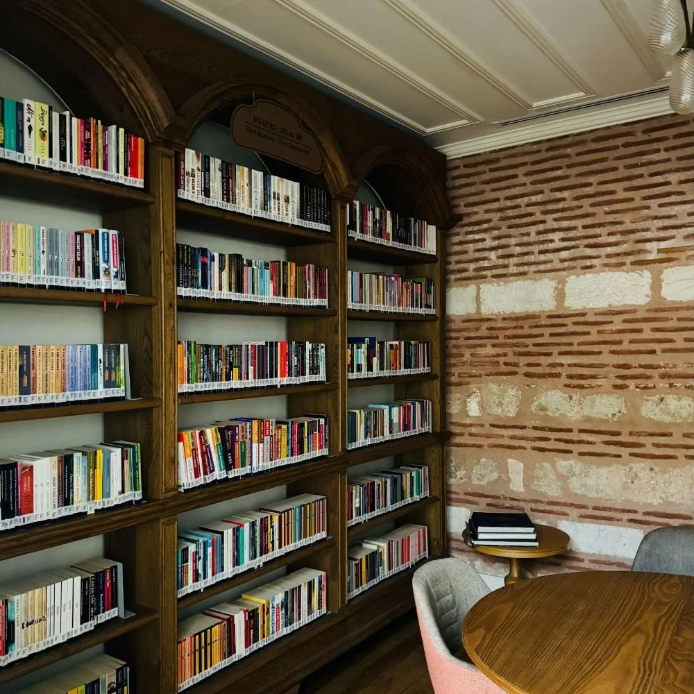 Bookshelves filled with colorful books in a cozy reading room with a brick wall, a round wooden table, and chairs.
