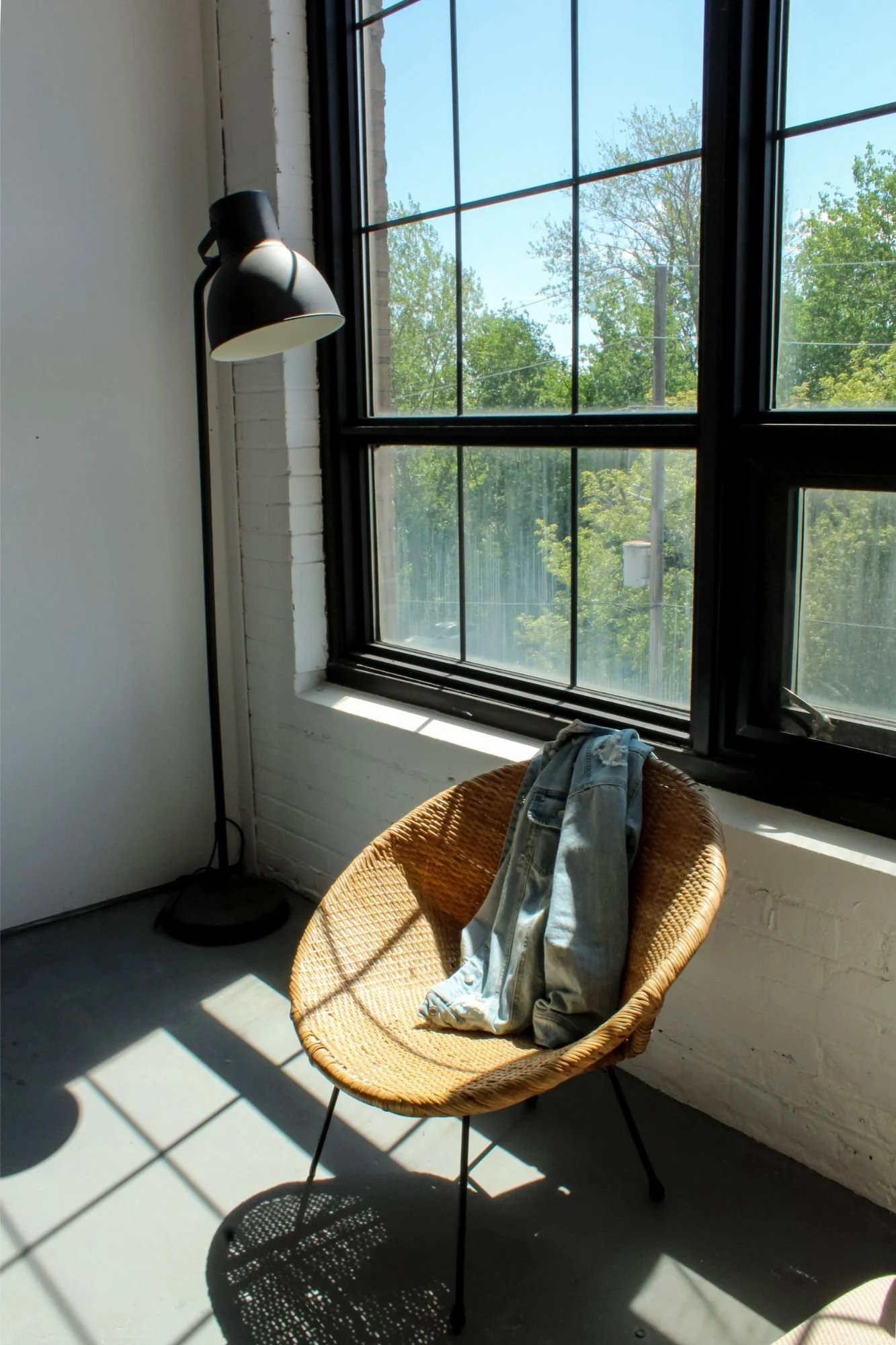 Sunlit room with a wicker chair holding a pair of jeans, a black floor lamp, and large window showing green trees outside.